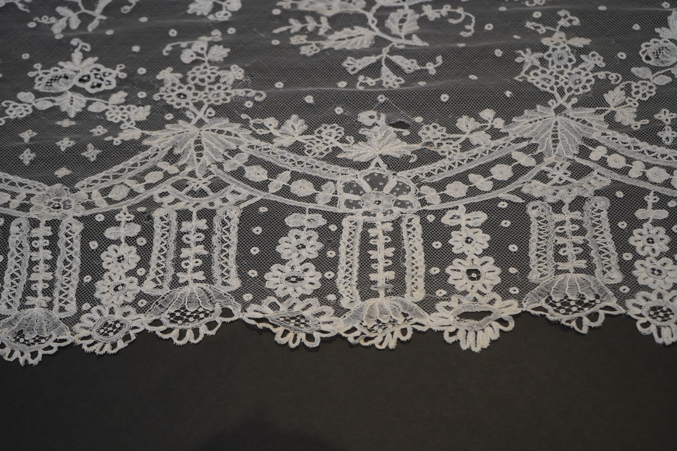 A deep elaborately designed flounce of mid 19th century Brussels bobbin appliquéd lace on net, with finished lace ends, worked with an intricate floral border below large floral cartouches framed in similar motifs and al